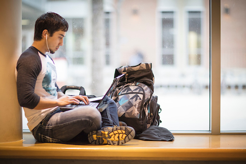 male presenting student wearing headphones sitting crosslegged and working on a laptop
